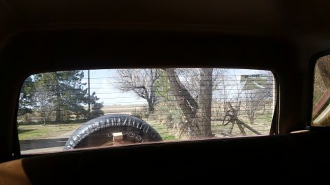 1985 Ford Bronco - View from inside the vehicle looking out the rear window, with a spare tire visible and a rural scene beyond.
