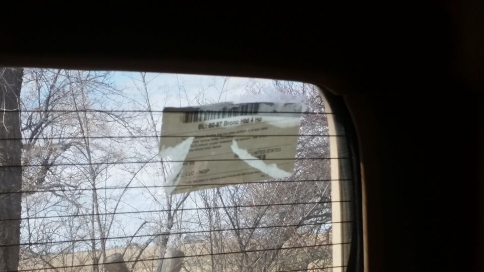 Sticker on a rear window reads 'BL-80-47 Bronco 1984 1985'. Trees and sky visible through the window
