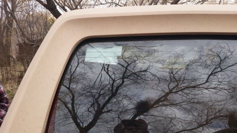 1985 Ford Bronco - The rear window of a beige SUV shows reflections of trees and a watermark from Supermotors.net.