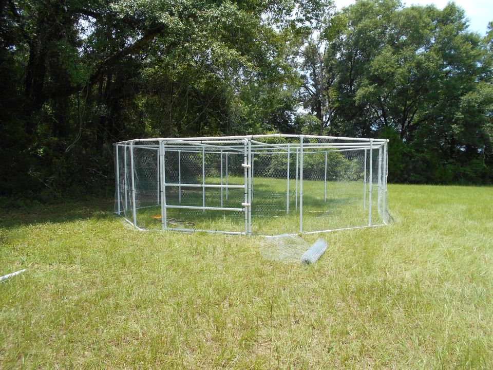 Octagonal wire mesh enclosure with metal frame and double doors, set in a grassy field with trees in the background. A roll of wire mesh lies on the grass