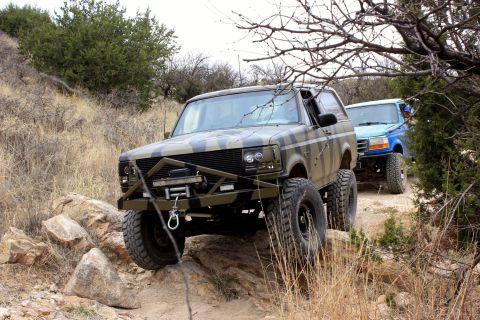 1996 Ford Bronco - Two off-road vehicles, a camouflaged Bronco and a blue truck, navigate a dirt path with rocky inclines.