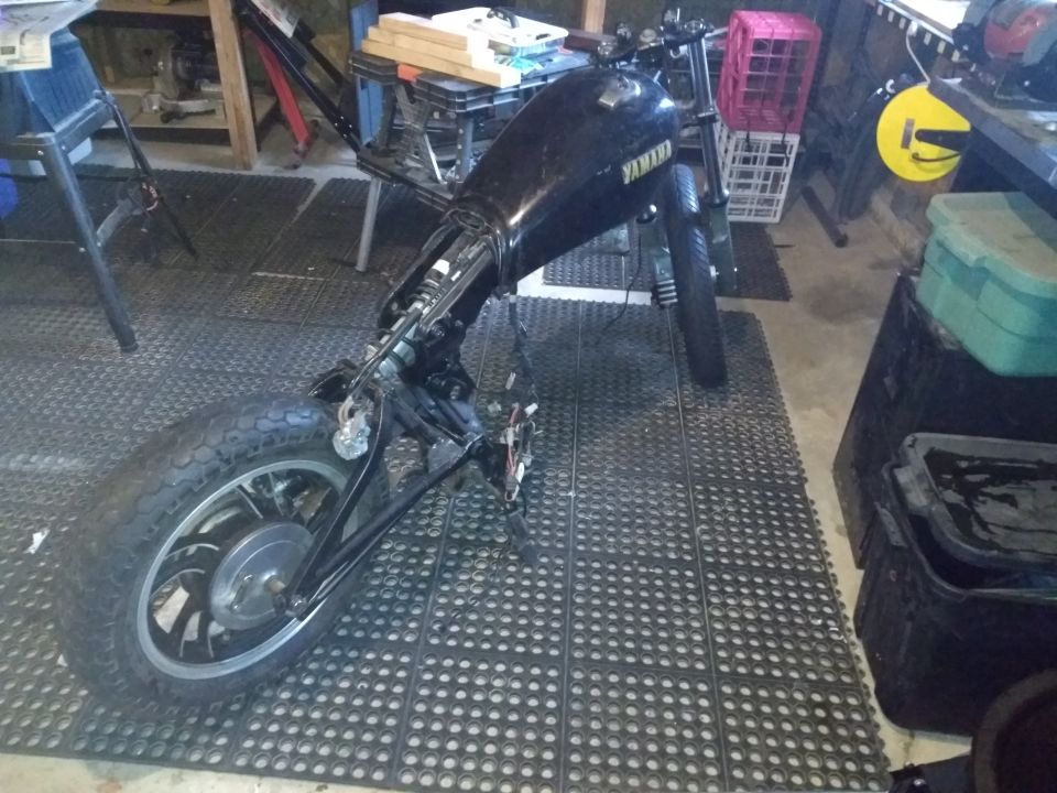 Partially disassembled black Yamaha motorcycle on garage floor mats, rear quarter view with exposed wiring and frame.
