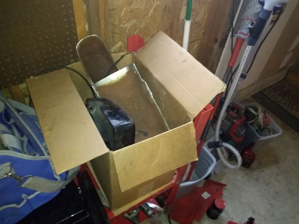 Open cardboard box in a garage holding black motorcycle parts and wiring, sitting on red shop equipment.