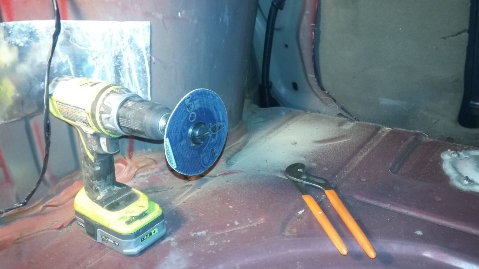 Drill with blue cutting disc and orange-handled pliers on rusty car floor. Metal panel behind drill