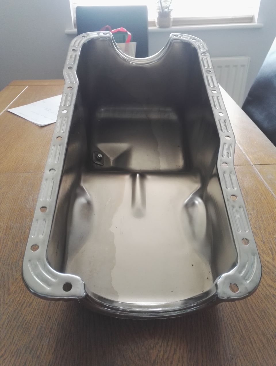 Shiny metal oil pan with mounting holes on a wooden table.