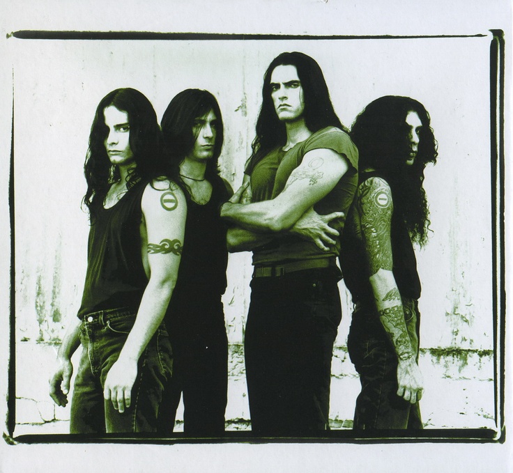 Four men with long dark hair and tattoos standing in front of a white wall.