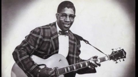 1967 Ford Custom Wagon - A black and white studio portrait of a man playing a guitar. He is wearing glasses and a plaid suit with a bow tie.