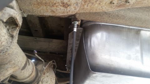 1985 Ford Bronco - Underneath view of a vehicle's fuel tank and rear axle assembly, showing rusted metal components and a fuel line connected to the tank.