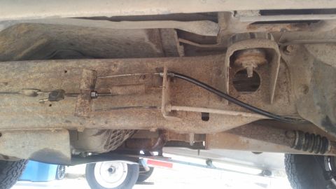 1985 Ford Bronco - The underside of a vehicle showing the rusty frame, suspension components, and brake cables. A black brake cable housing is visible running…