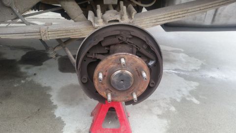 1985 Ford Bronco - A close-up view of the rear drum brake assembly of a vehicle, supported by a red jack stand. The brake drum is rusty and shows signs of wea…