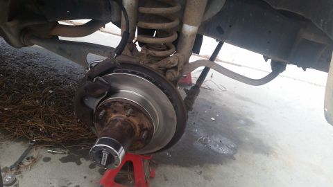 1985 Ford Bronco - A close-up view of the rear brake rotor and caliper assembly of a vehicle, supported by a red jack stand. Tools, including a wrench and soc…