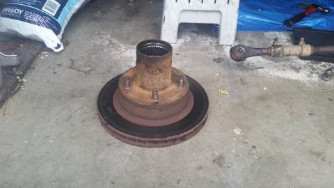 1985 Ford Bronco - A rusty brake rotor and hub assembly sits on a concrete surface, with tools and a bag of chemicals in the background.