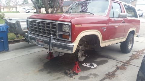 1985 Ford Bronco - The front end of a red Bronco is raised on jack stands, with brake components visible on the ground.