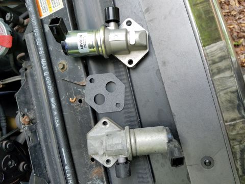 1995 Ford Bronco - Two idle air control valves and a gasket are laid out on a dark surface, possibly in an engine bay.
