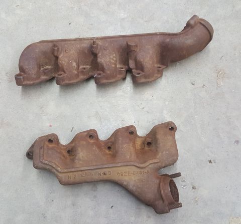 1973 Ford Mustang - Two old, rusty exhaust manifolds are shown resting on a concrete surface. Part numbers are visible on the lower manifold.