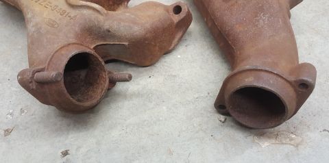 1973 Ford Mustang - Close-up of two aged exhaust manifolds, one bearing a Ford logo and part number, resting on a concrete surface.