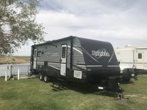 2018 Yugo GV - A travel trailer with 'Pioneer' branding is parked on a grassy area next to a body of water, with another RV visible in the background.