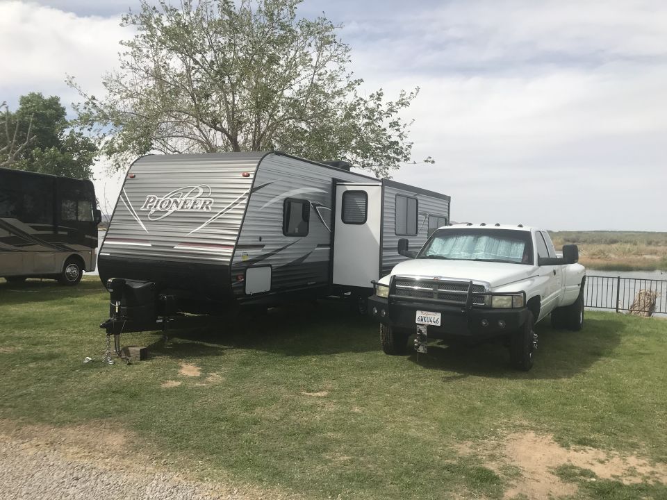 White pickup truck with a heavy-duty bumper towing a Pioneer travel trailer. Another RV is parked to the left