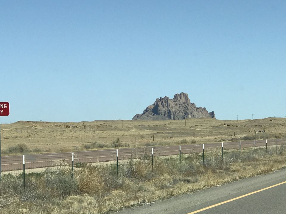 Large rock formation in a dry, grassy landscape under a clear blue sky. A road and fence are in the foreground. A red sign is visible on the left