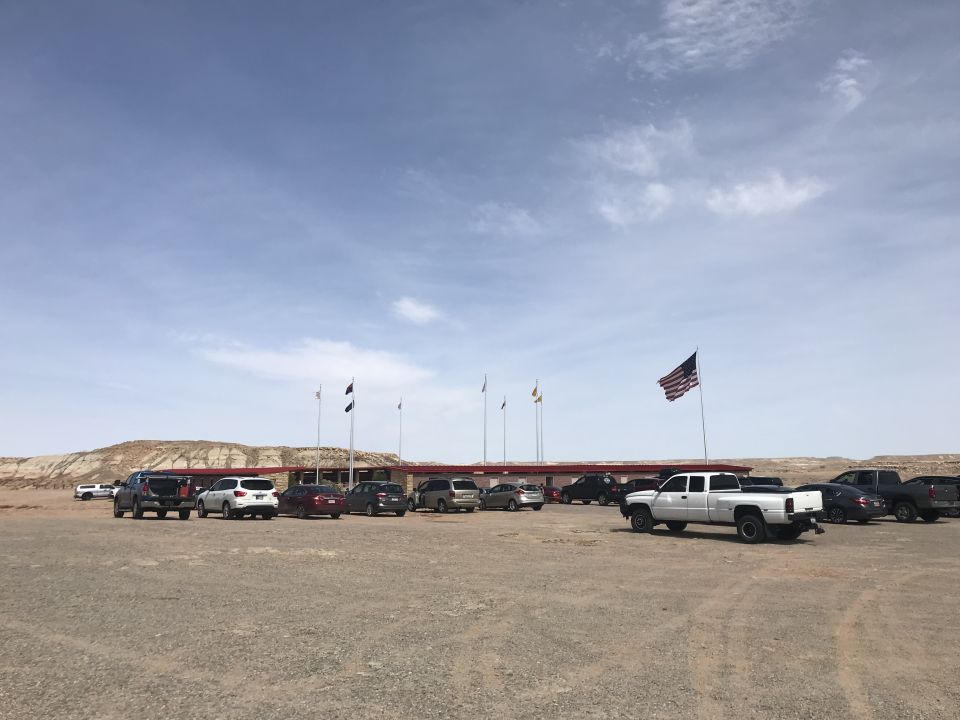 Parking lot with several cars and trucks in front of a building with flags, under a partly cloudy sky