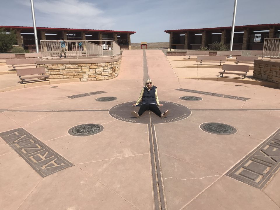 Person sitting on the ground at the Four Corners Monument, with markers for Arizona, Utah, Colorado, and New Mexico visible