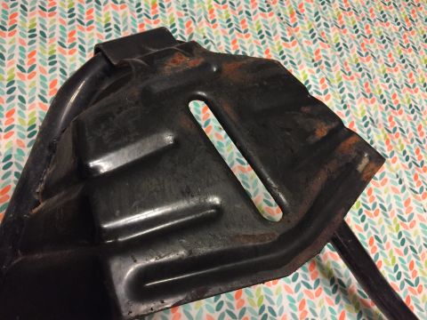 1967 Ford Custom Wagon - A black metal part with visible rust is shown against a colorful, patterned background. The part has a curved edge and a rectangular cutout.