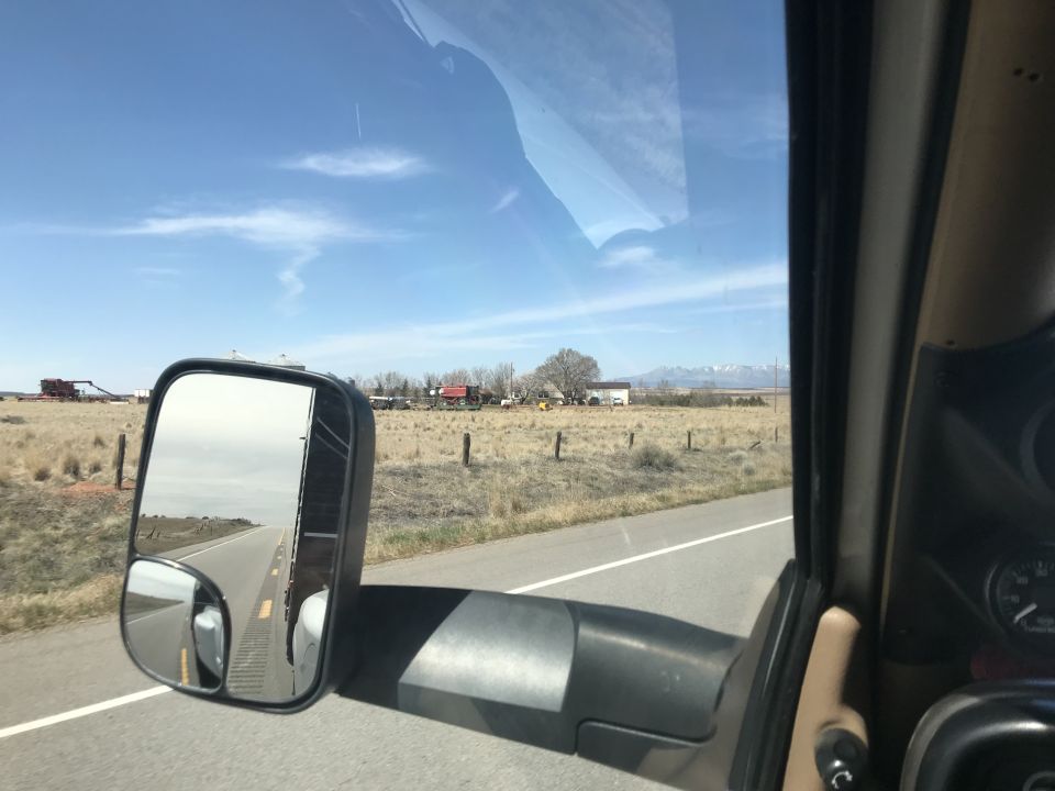 View from inside a vehicle showing a road and trailer in the side mirror, with a rural landscape outside.