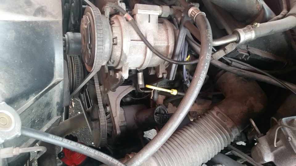 Engine bay with A/C compressor, belts, hoses, and yellow dipstick.