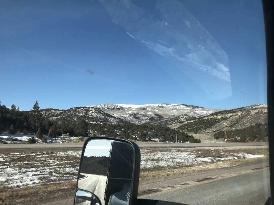 Snowy mountain landscape seen through a vehicle windshield and side mirror reflecting a dirt road.