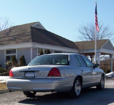 2002 Ford Crown Victoria - A silver 2002 Ford Crown Victoria is parked outside a building next to a flagpole flying the American flag.