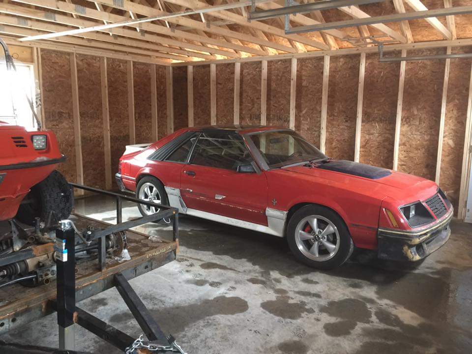 Red 1983 Ford Mustang coupe with black hood accent in a garage.