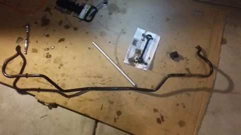 2000 Pontiac Grand Prix - A black metal sway bar is laid out on a cardboard surface with oil stains. Tools and a small packet of parts are also visible nearby.