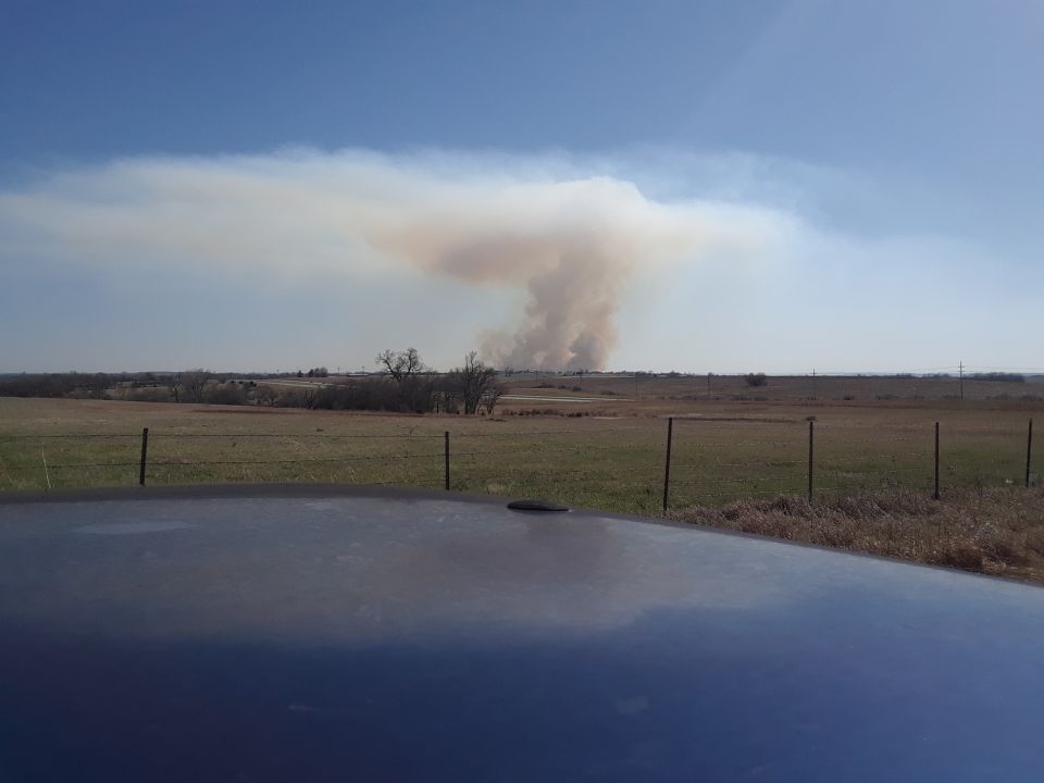 Smoke plume rising from a distant fire over a field, with a car hood in the foreground.