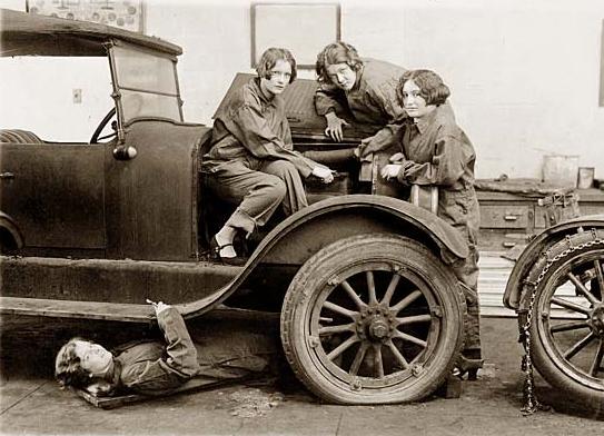 Three women with a vintage car, one underneath working on it.