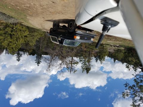 1968 Ford Bronco - A dark colored Ford Bronco is parked on a dirt road surrounded by pine trees and a bright blue sky with fluffy white clouds. The front of a…