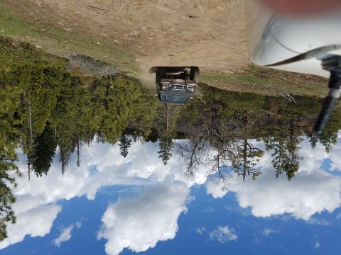1968 Ford Bronco - A dark colored SUV is driving on a dirt road surrounded by trees and a bright blue sky with white clouds. The vehicle appears to be kicking…