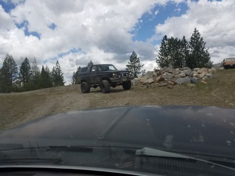 1968 Ford Bronco - A lifted black SUV with large tires is parked on a dirt road next to a pile of rocks and pine trees. The view is from inside another vehicl…