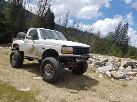 1968 Ford Bronco - A lifted white Ford F-250 pickup truck with large off-road tires is parked on a dirt road next to a pile of rocks and trees. The truck has…