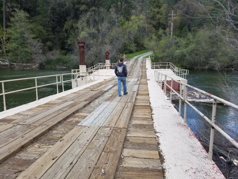 1968 Ford Bronco - A person walks across a wooden bridge over a river. The bridge has metal railings and appears to be an old railway bridge.