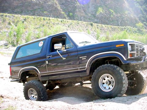 1988 Ford Bronco - This blue 1988 Ford Bronco is captured in a three-quarter front view while navigating a dirt terrain.
