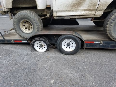1968 Ford Bronco - A flatbed trailer carries a dirty white truck, one of whose tires has blown out. The trailer has a single axle with two wheels, one of whic…
