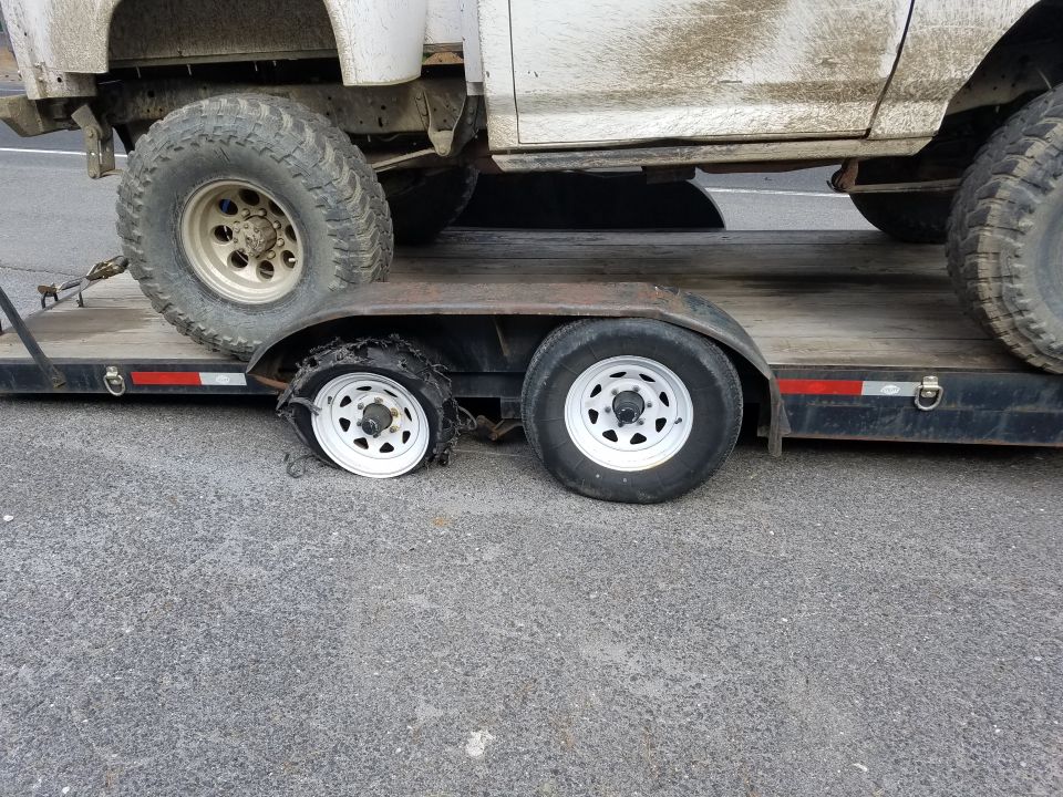 A flatbed trailer with a dirty white truck, showing a blown-out tire on the trailer.