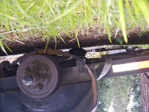 1968 Ford Bronco - A close-up view of a rusty trailer wheel and suspension system, with a ratchet strap attached to the trailer frame. Green grass and trees a…