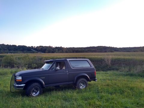 1996 Ford Bronco - A dark gray 1996 Ford Bronco is parked in a grassy field with a cornfield and trees in the background under a clear sky.