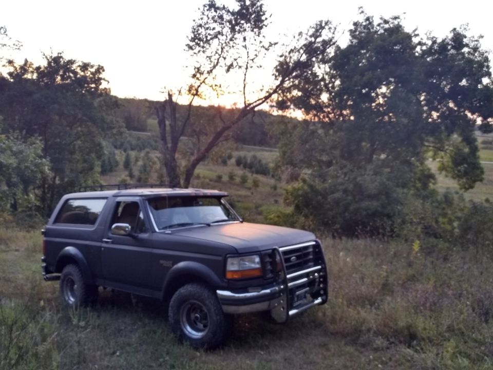 Dark 1996 Ford Bronco with brush guard and roof rack in a grassy field at dusk.