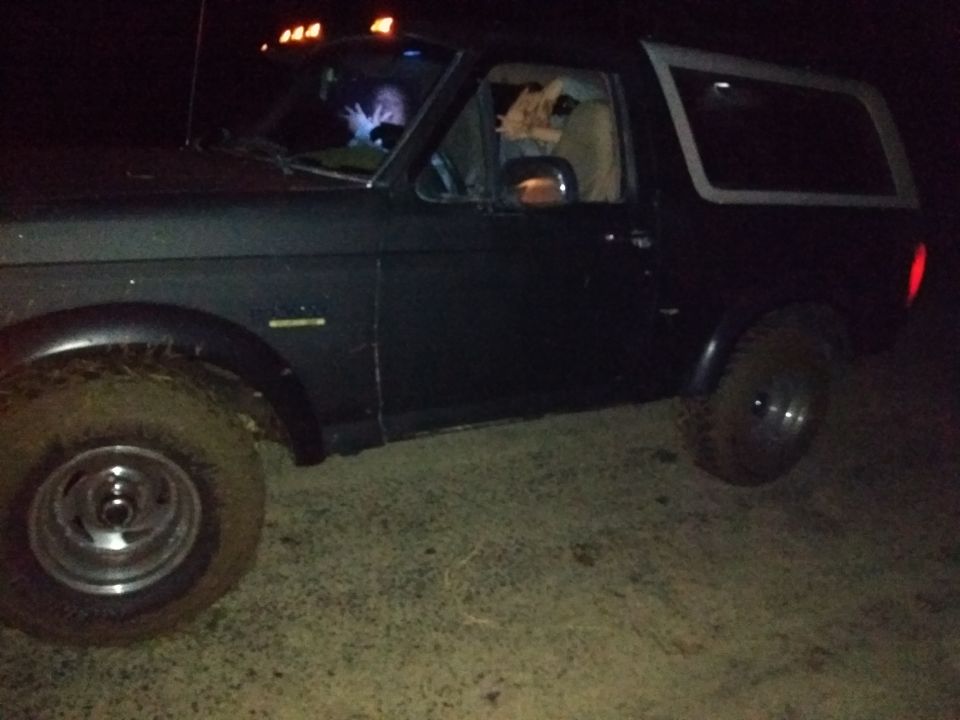 Dark-colored Ford Bronco with mud-splattered tires, viewed from the front-left at night
