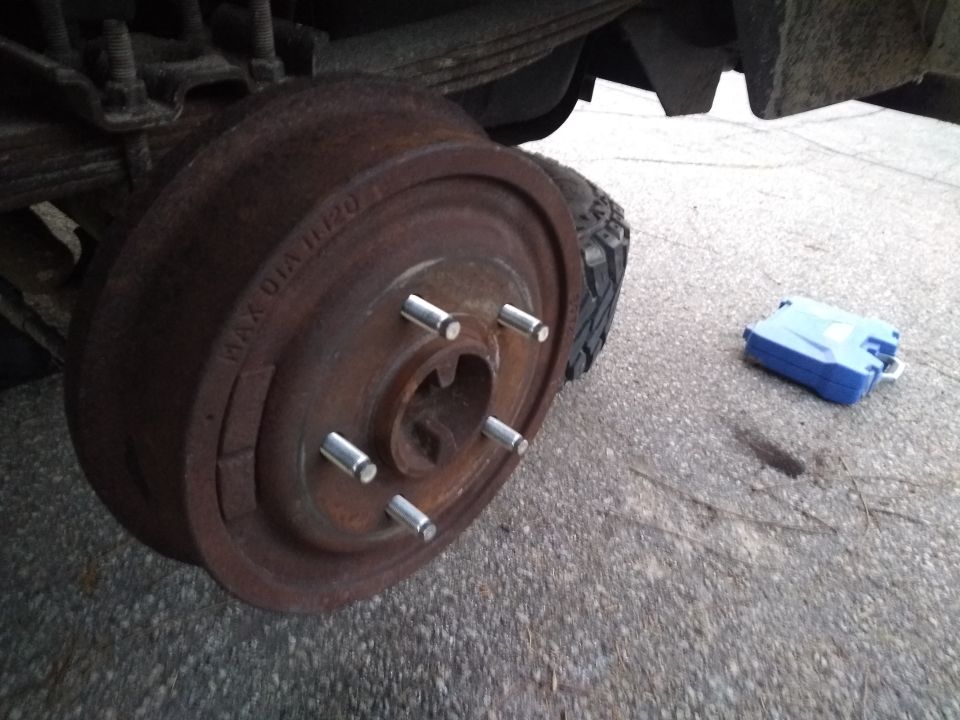 Rusty brake drum with new lug nuts on a vehicle, blue tool case on asphalt.