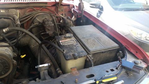 1985 Ford Bronco - Engine bay of an older Ford truck showing the battery, air intake tubing and wiring beside a red inner fender.
