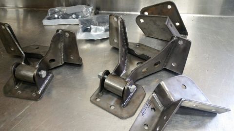 1979 Ford Bronco - Several custom-fabricated steel brackets with roller bearings are laid out on a stainless steel workbench. Some brackets have part numbers…