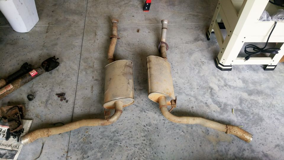 Two used exhaust pipes with oval mufflers lying on a concrete garage floor next to a driveshaft and bolts